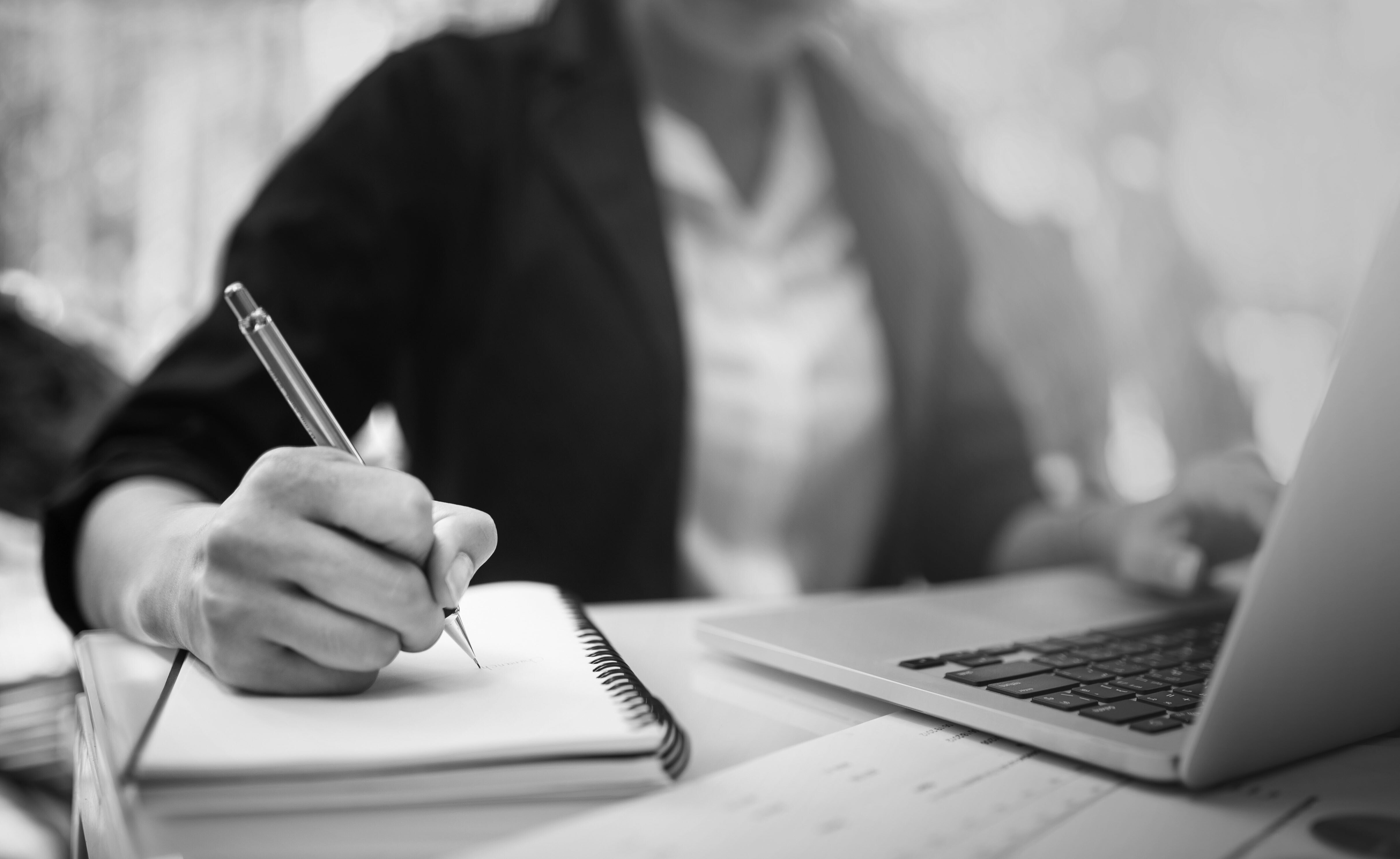 A black-and-white image of a person writing on a notepad with a pen near a laptop.